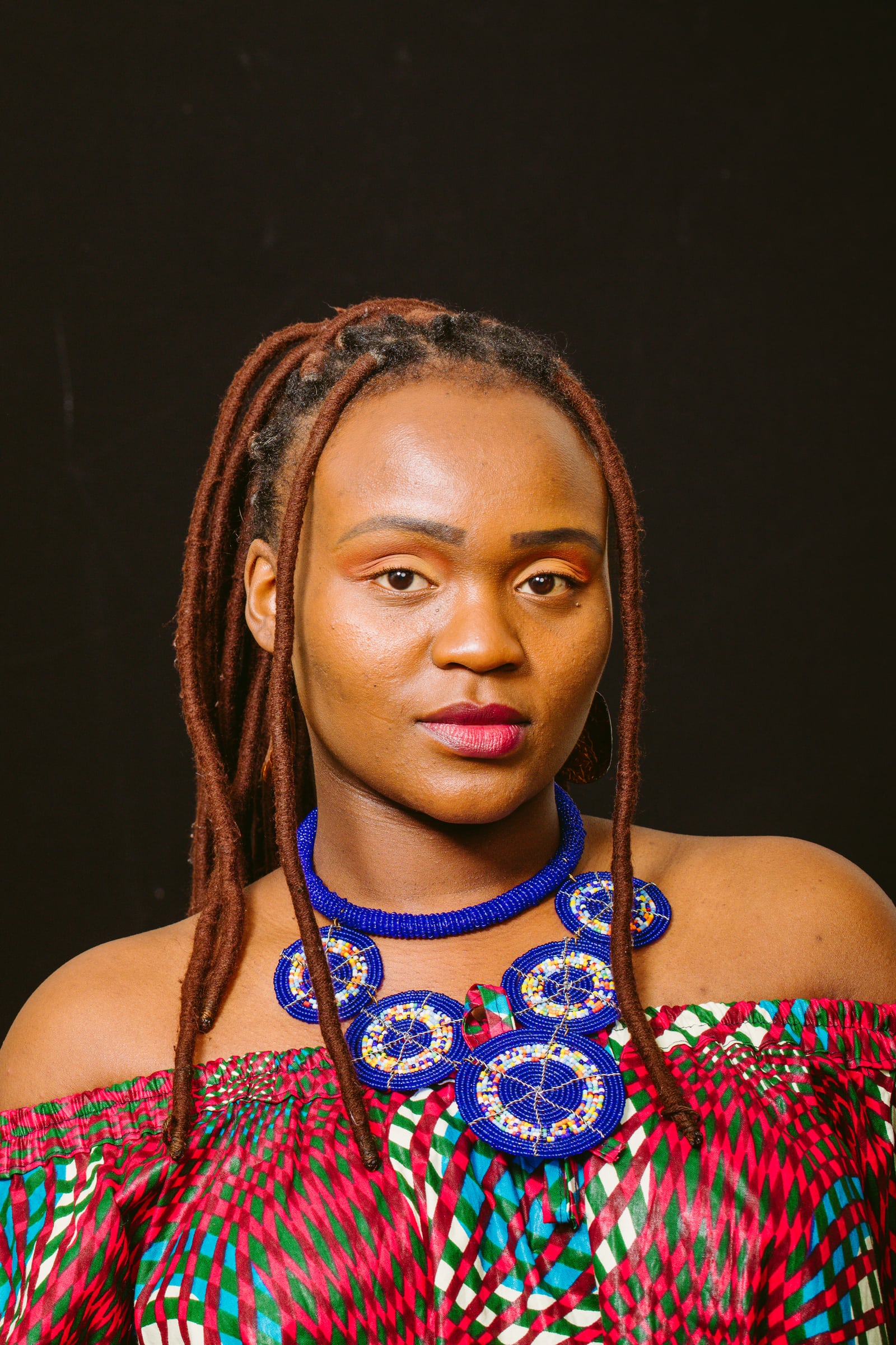 Woman braids + blue statement necklace, dark bg, elite cultural portrait. Photograph by Samuel Mfinanga for Mfinanga Photography + Motion.